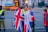 A group of people are gathered near a fenced-off area, with some wearing jackets featuring the Union Jack. Security personnel in high-visibility vests are present, and a large vehicle with a blue cover is visible in the background.
