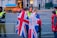 A group of people are gathered near a fenced-off area, with some wearing jackets featuring the Union Jack. Security personnel in high-visibility vests are present, and a large vehicle with a blue cover is visible in the background.