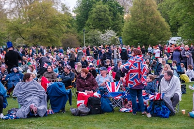 A large crowd of people gathered on a green lawn, many dressed in clothing featuring the Union Jack flag. The people are sitting on camping chairs and wrapped in blankets, suggesting a festive outdoor event. Trees and lush greenery surround the area, indicating a park-like setting.