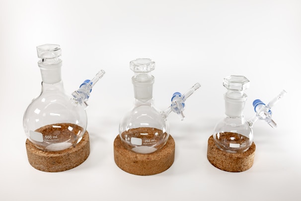 Three clear glass laboratory flasks of different sizes are placed in a row on circular cork bases. Each flask has a side arm with a blue and transparent connector.