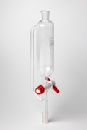 A clear glass laboratory separatory funnel with a cylindrical main body and a stopcock featuring red and white knobs. The apparatus is used for separating liquid mixtures.