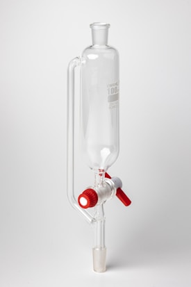 A clear glass laboratory separatory funnel with a cylindrical main body and a stopcock featuring red and white knobs. The apparatus is used for separating liquid mixtures.
