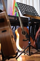 A wooden acoustic guitar rests against a stand next to a Yamaha keyboard and a music stand with sheet music. Various colorful objects are in the background, adding vibrancy to the scene. The lighting casts a warm glow on the instruments, enhancing the cozy ambiance.