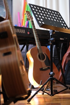 A wooden acoustic guitar rests against a stand next to a Yamaha keyboard and a music stand with sheet music. Various colorful objects are in the background, adding vibrancy to the scene. The lighting casts a warm glow on the instruments, enhancing the cozy ambiance.