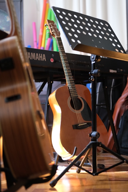 A wooden acoustic guitar rests against a stand next to a Yamaha keyboard and a music stand with sheet music. Various colorful objects are in the background, adding vibrancy to the scene. The lighting casts a warm glow on the instruments, enhancing the cozy ambiance.