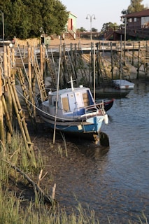 A small wooden boat with a blue and white cabin is moored in shallow water alongside a dock made of wooden posts and ropes. The surrounding area features grassy banks and rustic buildings with stairs leading down to the waterfront, creating a serene and rustic harbor scene.