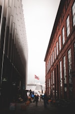 A narrow street between two buildings, with a modern gray structure on the left and a red brick building on the right. A Norwegian flag is seen in the background above a large cruise ship. People are walking along the street, and bicycles are parked on the right sidewalk.