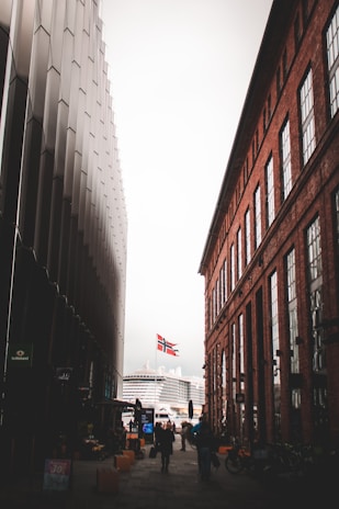 A narrow street between two buildings, with a modern gray structure on the left and a red brick building on the right. A Norwegian flag is seen in the background above a large cruise ship. People are walking along the street, and bicycles are parked on the right sidewalk.