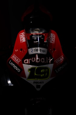 A dark, dramatic image focusing on the front of a racing motorcycle with bright red and white livery. The number 19 is prominently displayed in the center, surrounded by various sponsor logos such as 'aruba.it', 'Lenovo', 'Fiamm', and 'Stulz'. The lighting highlights the aggressive design and aerodynamic shape of the bike.