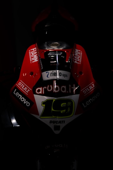 A dark, dramatic image focusing on the front of a racing motorcycle with bright red and white livery. The number 19 is prominently displayed in the center, surrounded by various sponsor logos such as 'aruba.it', 'Lenovo', 'Fiamm', and 'Stulz'. The lighting highlights the aggressive design and aerodynamic shape of the bike.