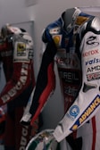 Two motorcycle racing suits are displayed side by side, adorned with various sponsor logos and designs. The suit in the foreground features logos such as Xerox, AMD, and Shell, and has a color scheme of red, blue, and white with intricate graphics. A helmet with a striking design can be seen at the bottom of the display.