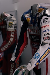 Two motorcycle racing suits are displayed side by side, adorned with various sponsor logos and designs. The suit in the foreground features logos such as Xerox, AMD, and Shell, and has a color scheme of red, blue, and white with intricate graphics. A helmet with a striking design can be seen at the bottom of the display.