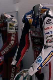 Two motorcycle racing suits are displayed side by side, adorned with various sponsor logos and designs. The suit in the foreground features logos such as Xerox, AMD, and Shell, and has a color scheme of red, blue, and white with intricate graphics. A helmet with a striking design can be seen at the bottom of the display.