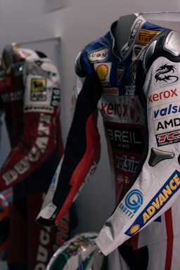 Two motorcycle racing suits are displayed side by side, adorned with various sponsor logos and designs. The suit in the foreground features logos such as Xerox, AMD, and Shell, and has a color scheme of red, blue, and white with intricate graphics. A helmet with a striking design can be seen at the bottom of the display.