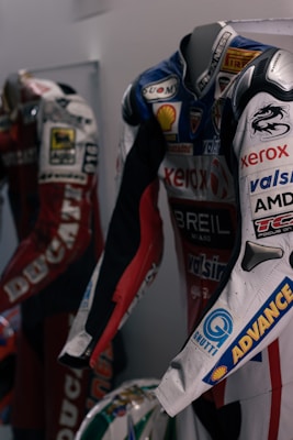 Two motorcycle racing suits are displayed side by side, adorned with various sponsor logos and designs. The suit in the foreground features logos such as Xerox, AMD, and Shell, and has a color scheme of red, blue, and white with intricate graphics. A helmet with a striking design can be seen at the bottom of the display.
