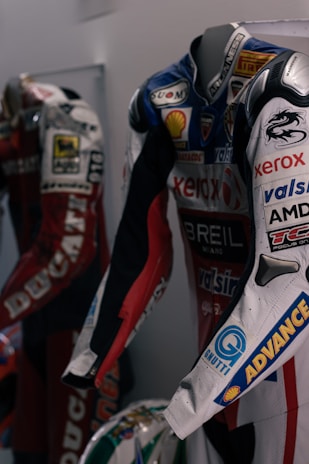Two motorcycle racing suits are displayed side by side, adorned with various sponsor logos and designs. The suit in the foreground features logos such as Xerox, AMD, and Shell, and has a color scheme of red, blue, and white with intricate graphics. A helmet with a striking design can be seen at the bottom of the display.