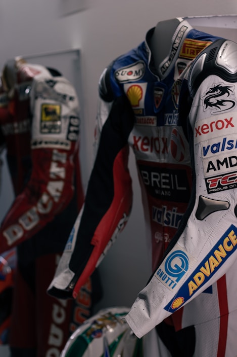 Two motorcycle racing suits are displayed side by side, adorned with various sponsor logos and designs. The suit in the foreground features logos such as Xerox, AMD, and Shell, and has a color scheme of red, blue, and white with intricate graphics. A helmet with a striking design can be seen at the bottom of the display.