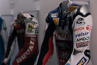 Three racing suits are displayed on mannequins. The suit in the foreground has several sponsor logos, including Xerox, Shell, and AMD, and features a prominent dragon design on the upper arm. The color scheme is predominantly white, red, and black with some blue accents. The suit behind it is red and white with the word 'DUCATI' visible. The display is in an indoor environment, likely a showroom or a museum.