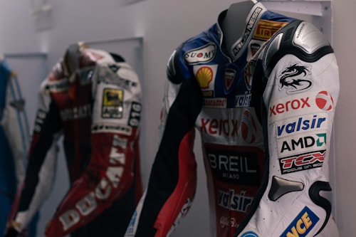Three racing suits are displayed on mannequins. The suit in the foreground has several sponsor logos, including Xerox, Shell, and AMD, and features a prominent dragon design on the upper arm. The color scheme is predominantly white, red, and black with some blue accents. The suit behind it is red and white with the word 'DUCATI' visible. The display is in an indoor environment, likely a showroom or a museum.