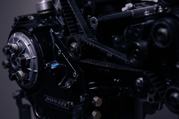 A close-up view of a mechanical component, likely part of an engine, showcasing intricate details such as gears, bolts, and belts. The image features the logo 'Ducati' prominently displayed on the machinery.