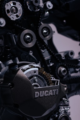 Close-up view of intricate parts of a mechanical engine. Prominent features include gears, pulleys, chains, and a casing with the word 'DUCATI' embossed on it. The image captures the precision and complexity of the engine's components.