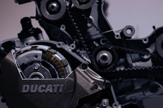 A close-up view of a motorcycle engine, showcasing intricate mechanical parts like gears, belts, and bolts with a visible 'DUCATI' branding. The image highlights the metallic textures and complexity of the engine components.