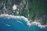 An aerial view of a coastline featuring rugged terrain with dense green forests meeting the rocky shore. The clear blue ocean contrasts with the land, and several boats are visible near the shoreline, creating trails in the water. A rectangular structure is noticeable amidst the greenery.