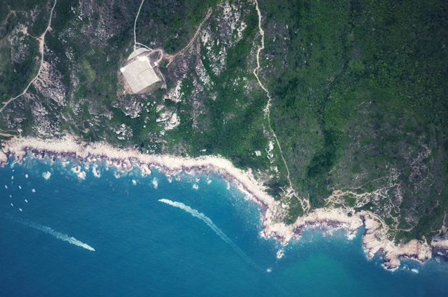 An aerial view of a coastline featuring rugged terrain with dense green forests meeting the rocky shore. The clear blue ocean contrasts with the land, and several boats are visible near the shoreline, creating trails in the water. A rectangular structure is noticeable amidst the greenery.