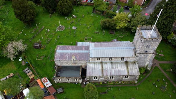 An aerial view of a stone church with a tower and a flagpole, surrounded by a cemetery and lush green grass. The image features numerous gravestones scattered across the grounds and trees lining the perimeter. The church's roof is made of metal panels, some of which appear weathered.