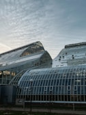 A large glasshouse structure with a complex design, featuring multiple glass panels and a curved roof in a modern architectural style against a partly cloudy sky.