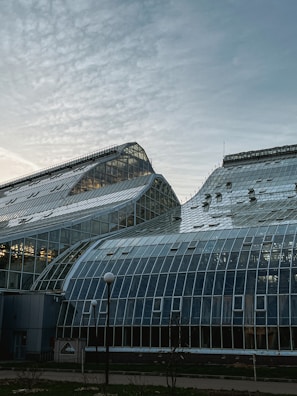 A large glasshouse structure with a complex design, featuring multiple glass panels and a curved roof in a modern architectural style against a partly cloudy sky.