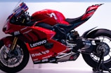 A sleek, red racing motorcycle featuring several sponsor logos such as Lenovo and NetApp. The bike has a streamlined design with a prominent windshield and visible mechanical components. Its rear swingarm and large rear tire indicate its high-performance capabilities.