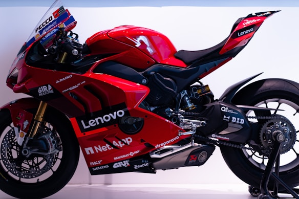 A sleek, red racing motorcycle featuring several sponsor logos such as Lenovo and NetApp. The bike has a streamlined design with a prominent windshield and visible mechanical components. Its rear swingarm and large rear tire indicate its high-performance capabilities.