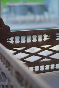 An intricately designed wooden bench with a lattice backrest and spindles, placed in an outdoor setting. The foreground is sharp, while the background is blurred, creating a depth of field effect with some greenery visible.