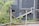 A sign labeled 'Assessment Centre' points to the right, surrounded by a metal fence. Behind the fence, there are plants and foliage, suggesting an outdoor setting. The background is dominated by grey panels, giving an industrial or construction site appearance.