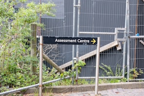 A sign labeled 'Assessment Centre' points to the right, surrounded by a metal fence. Behind the fence, there are plants and foliage, suggesting an outdoor setting. The background is dominated by grey panels, giving an industrial or construction site appearance.
