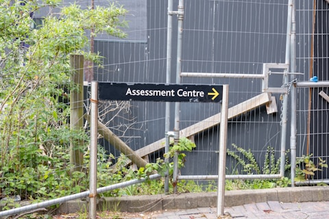 A sign labeled 'Assessment Centre' points to the right, surrounded by a metal fence. Behind the fence, there are plants and foliage, suggesting an outdoor setting. The background is dominated by grey panels, giving an industrial or construction site appearance.