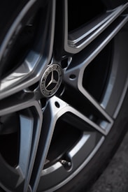 A close-up view of a metallic car wheel featuring a silver, multi-spoke rim with a central logo emblem. The surface of the wheel appears polished and clean, capturing reflections of the surroundings.