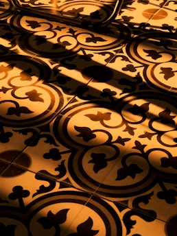 Ornate floor tiles with intricate floral and geometric patterns are illuminated by beams of warm, golden sunlight creating contrasting shadows.