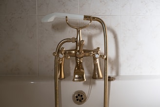 A vintage-style bathtub faucet with a gold finish features two separate knobs labeled 'H' and 'C' for hot and cold water. The faucet includes a handheld shower head attached by a flexible metal hose. The background consists of light, marbled tiles.
