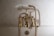 A vintage-style bathtub faucet with a gold finish features two separate knobs labeled 'H' and 'C' for hot and cold water. The faucet includes a handheld shower head attached by a flexible metal hose. The background consists of light, marbled tiles.