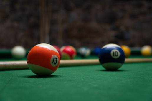 A close-up of billiard balls on a green pool table, including an orange-striped ball with the number 13 and a blue-striped ball with the number 10. A pool cue is resting on the table, and the background is blurred.