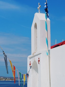 A white-walled church with a prominent cross at the top is adorned with colorful flags and a Greek flag. The church is located near a blue sea under a clear blue sky.