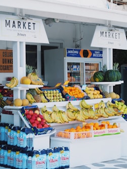 A small outdoor market stand displays a variety of fresh fruits such as bananas, oranges, pineapples, watermelons, and melons. Shelves are neatly organized with colorful produce set against a white backdrop. In the background, there is a refrigerator containing bottled and canned drinks, with signs above advertising 'Market' and 'Craft Beers'.
