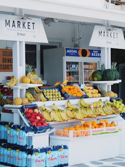 A small outdoor market stand displays a variety of fresh fruits such as bananas, oranges, pineapples, watermelons, and melons. Shelves are neatly organized with colorful produce set against a white backdrop. In the background, there is a refrigerator containing bottled and canned drinks, with signs above advertising 'Market' and 'Craft Beers'.