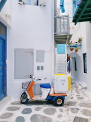 A narrow alley features a white building with blue accents, characterized by a small balcony and several potted plants. A scooter with a storage box on the back is parked in the alley on a stone pathway. The walls and doors are painted in bright white and vibrant blue, with colorful flowers adding contrast.