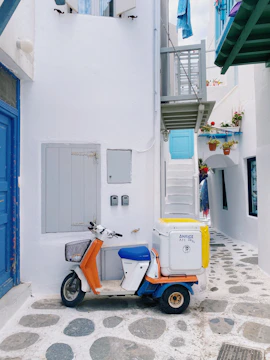 A narrow alley features a white building with blue accents, characterized by a small balcony and several potted plants. A scooter with a storage box on the back is parked in the alley on a stone pathway. The walls and doors are painted in bright white and vibrant blue, with colorful flowers adding contrast.