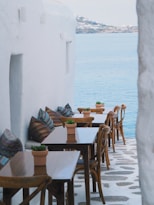 A serene seaside caf&eacute; with wooden tables and chairs lining a narrow pathway adjacent to a whitewashed building. Colorful cushions and small potted plants decorate the seating area, while the calm blue sea stretches out under a clear sky in the background.
