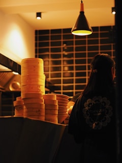 A dimly lit restaurant kitchen with stacks of bamboo steamers on a counter. A person is standing with their back to the camera, wearing a uniform with 'Tokyo Joe' written on it. The lighting is warm, highlighting the steamers and creating a cozy atmosphere.