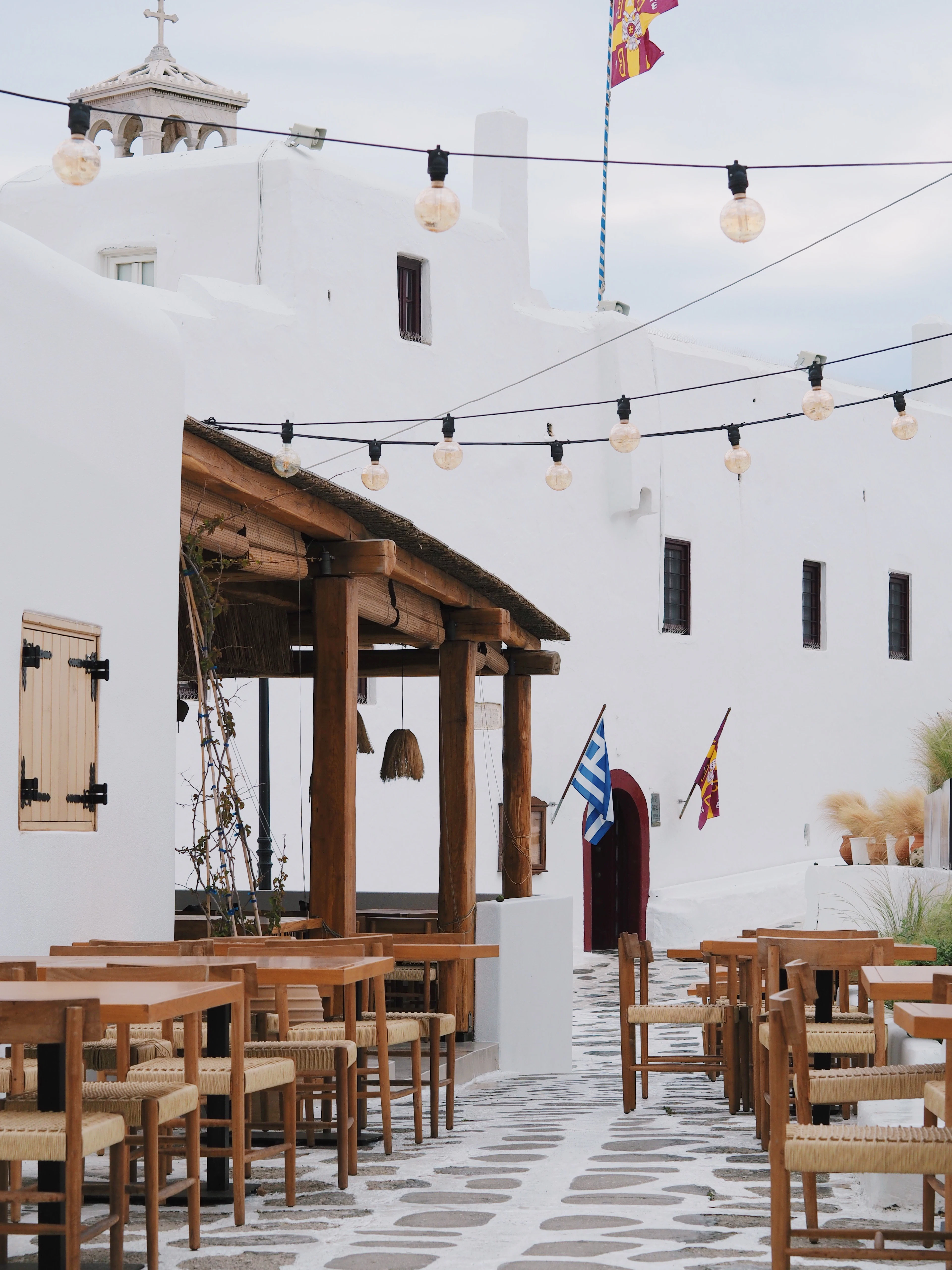 The quiet square at Ano Mera on Mykonos: empty wooden taverna tables and rush-seated chairs under a string of festoon lights, beside a climbing-plant pergola and the whitewashed walls and bell cote of the Panagia Tourliani monastery.